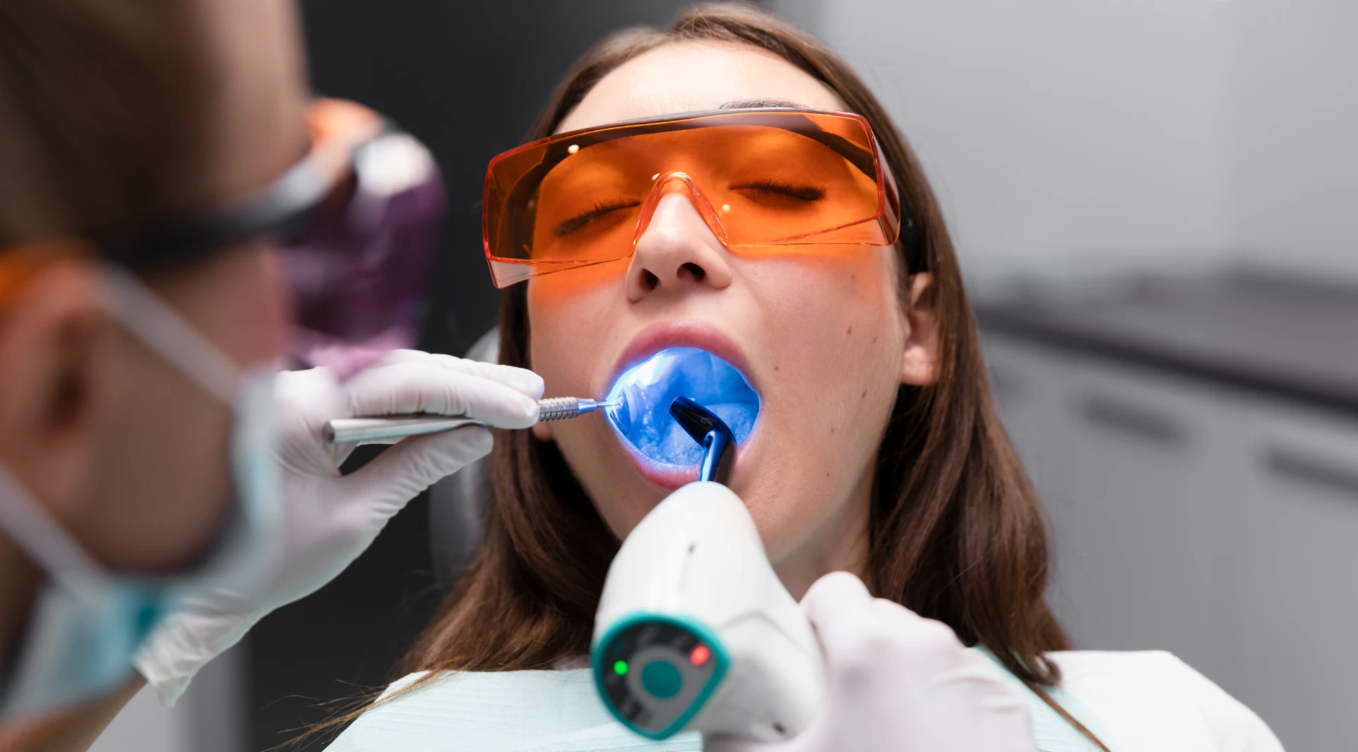 Pacientă cu ochelari de protecție portocalii în timpul unui tratament dentar cu lumină albastră UV, efectuat de medicul stomatolog
