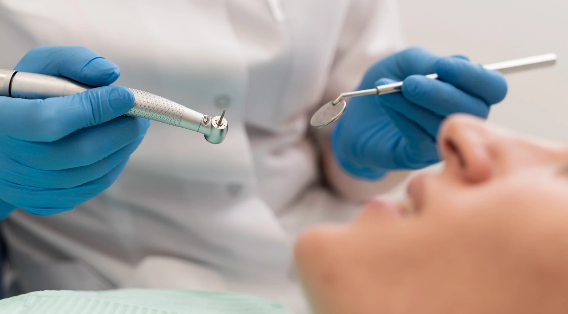 Mâini de medic stomatolog cu mănuși albastre ținând freză dentară și oglindă în timpul consultației unui pacient