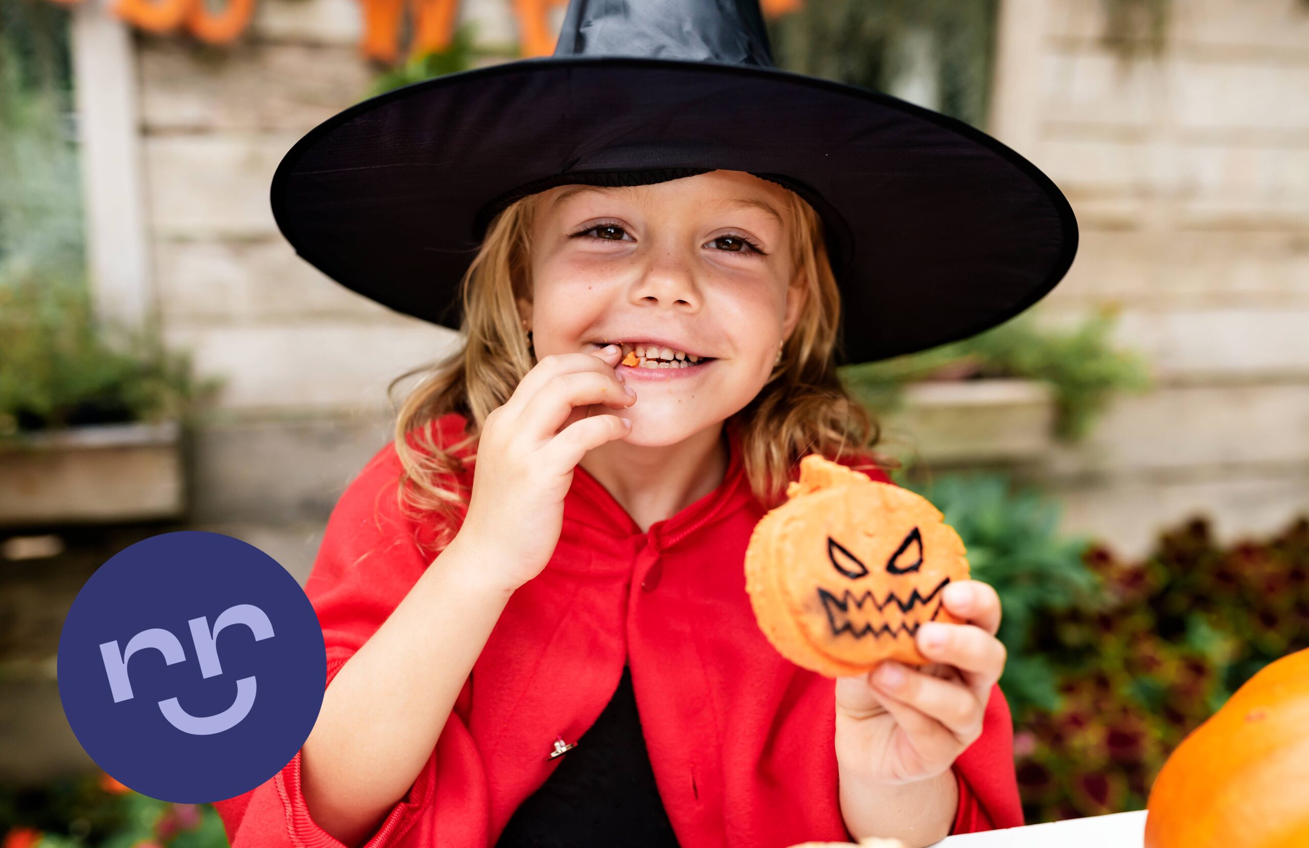 Fetiță în costum de Halloween, zâmbind și mâncând un biscuit în formă de dovleac